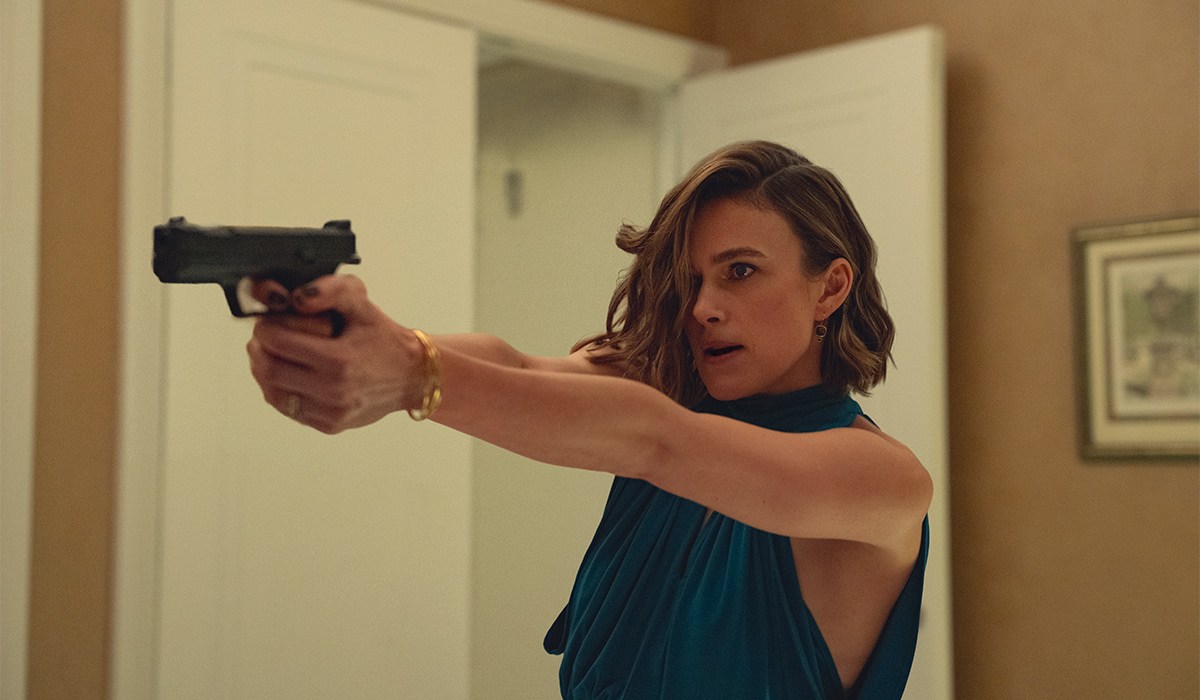 First look: Netflix drops trailer and release date for Black Doves season two 3 "Woman in blue dress aiming a handgun with both hands in a tense indoor setting, standing by a white door with a picture frame on the beige wall in the background."