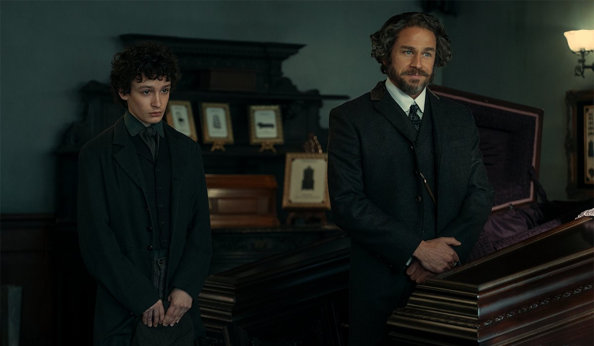 Dark and twisted new chapter of Monster series lands on Netflix 3 "Two men in dark, old-fashioned suits stand beside a polished wooden coffin in a dimly lit, ornate room decorated with framed photos, suggesting a 19th-century funeral or wake setting."